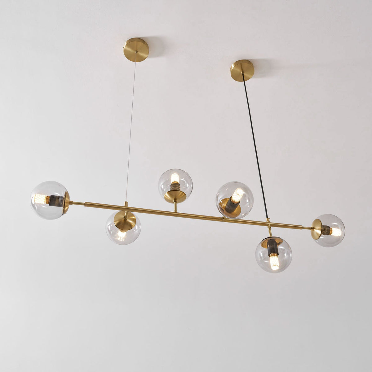 Seen at a slight angle against a white wall, this Clérachic pendant's lit clear orbs sit on a brass rod and two canopies.