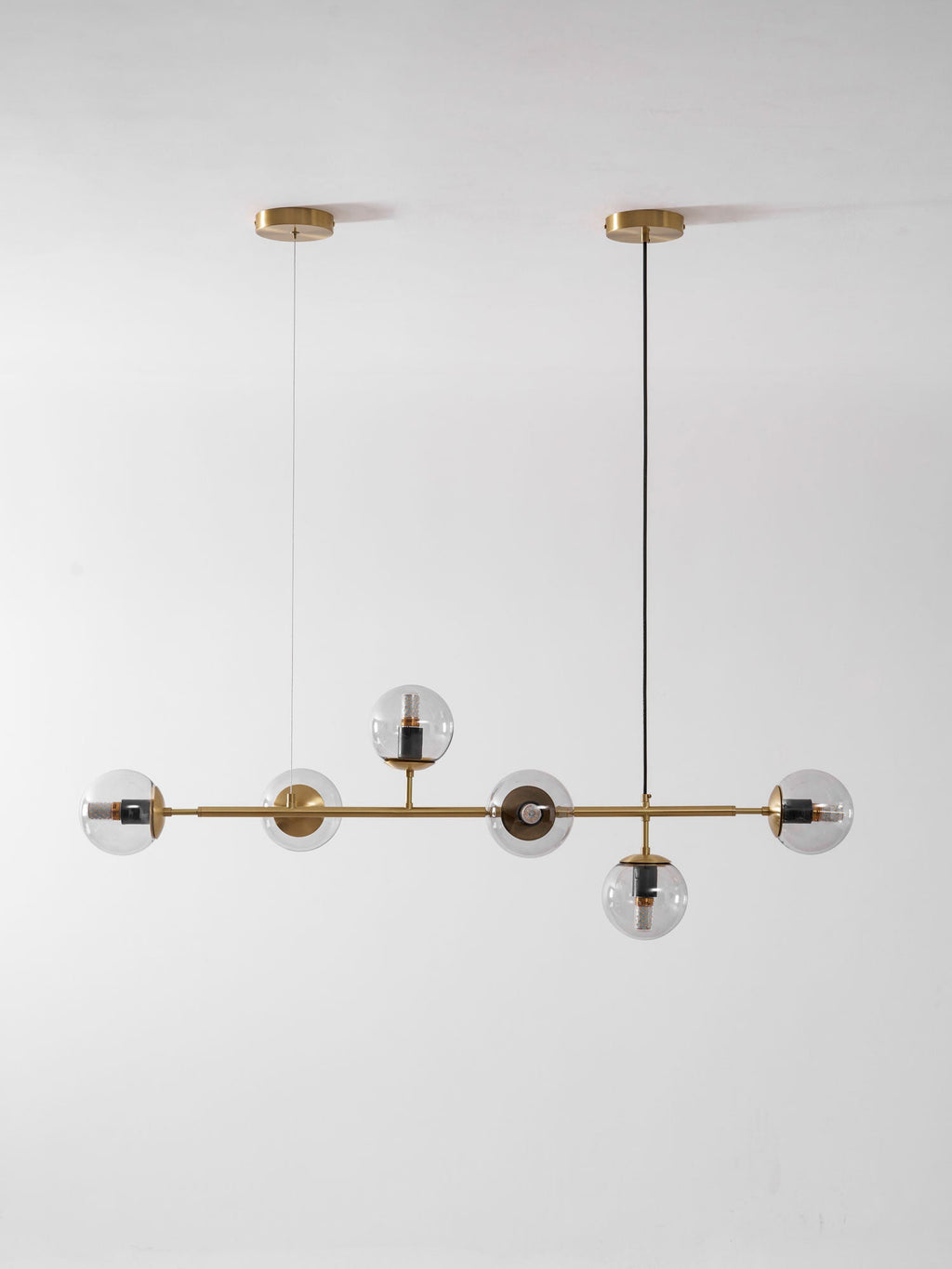 Smaller in frame, this Clérachic pendant hangs from two brass canopies, with six clear orbs on a brass rod on a pale wall.
