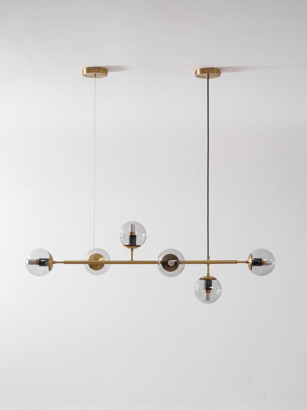 Smaller in frame, this Clérachic pendant hangs from two brass canopies, with six clear orbs on a brass rod on a pale wall.