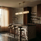 Clérachic gold-banded opal tube pendant above a stone-clad kitchen island with three bouclé stools and a concrete wall.