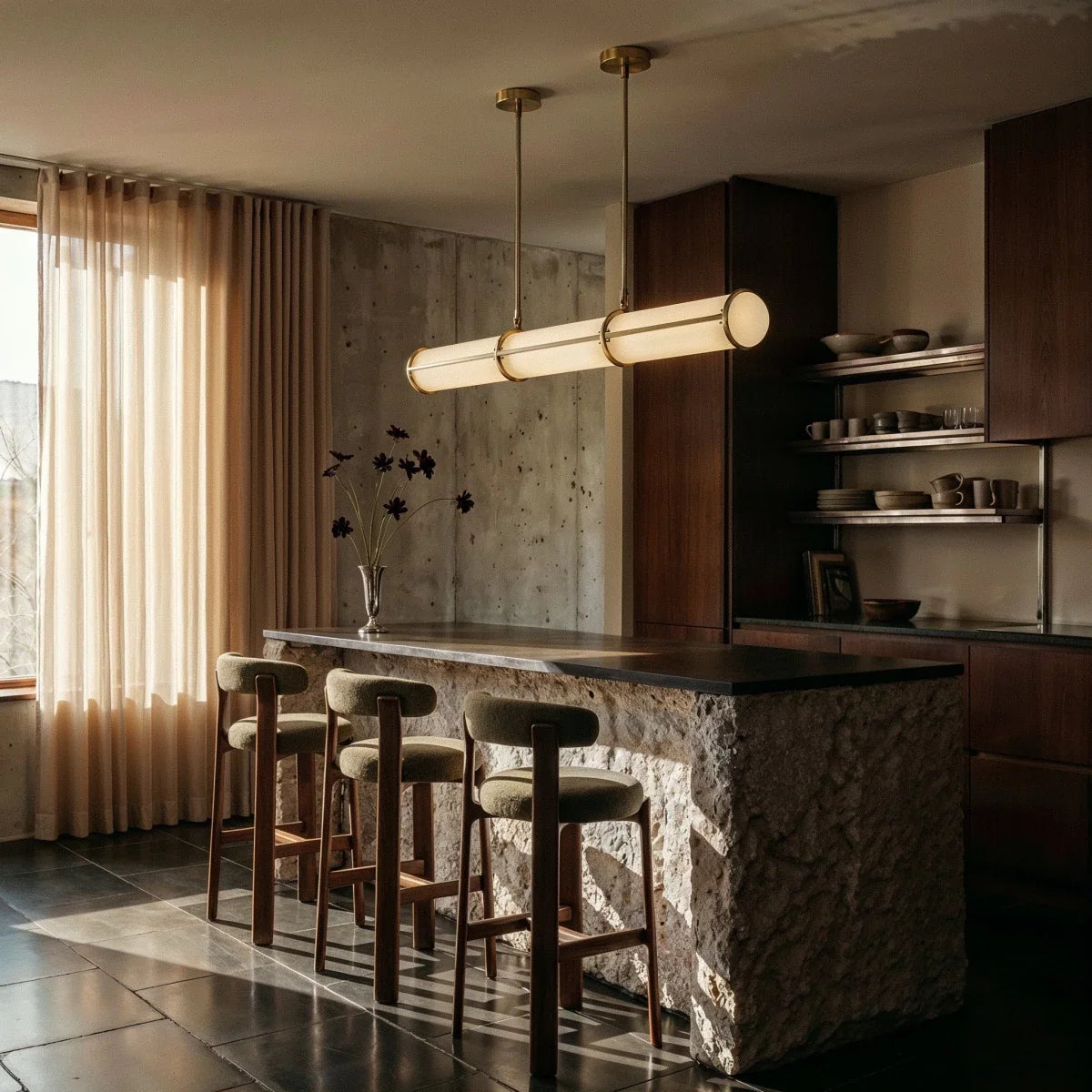 Clérachic gold-banded opal tube pendant above a stone-clad kitchen island with three bouclé stools and a concrete wall.