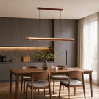 Over a walnut dining table with grey-cushioned chairs, a Clérachic linear pendant glows in front of grey kitchen cabinets.