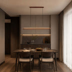 A Clérachic linear pendant glows over a dark dining table, in front of a tall cabinet with a lit shelf below.