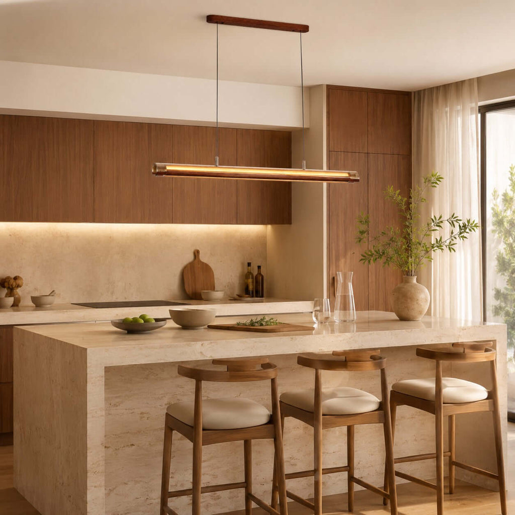 This Clérachic linear pendant lights a kitchen with a stone island, cream-cushioned stools and warm wood cabinets above.