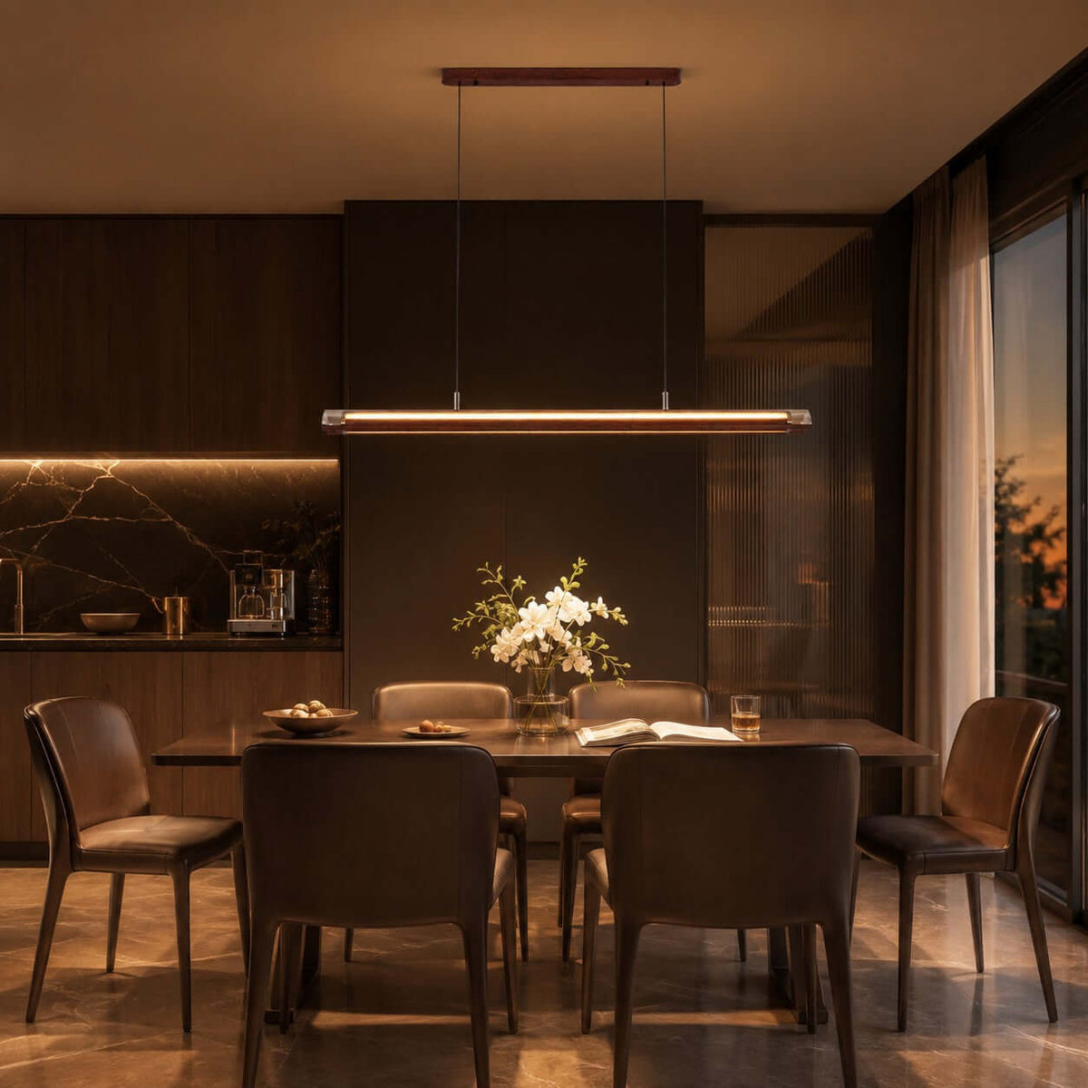 At dusk, a Clérachic linear pendant glows above a dark dining table with leather chairs, in a room with a marble backsplash.