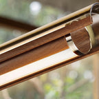 Tight view of the walnut casing, the metal ring and the diffuser strip of this Clérachic linear pendant, with a blurred garden behind.