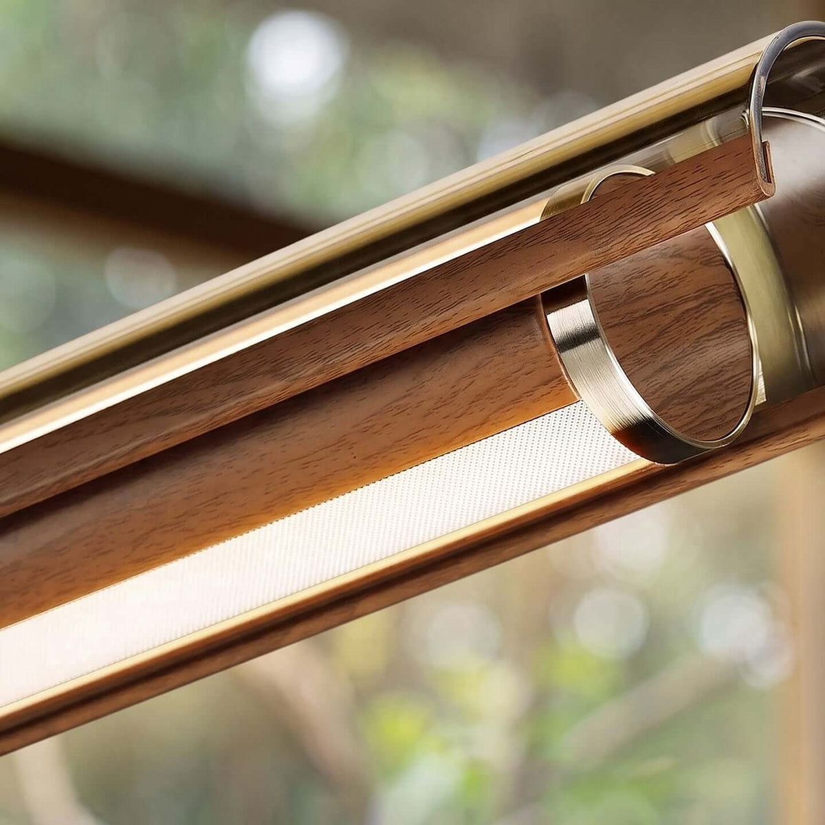 Tight view of the walnut casing, the metal ring and the diffuser strip of this Clérachic linear pendant, with a blurred garden behind.