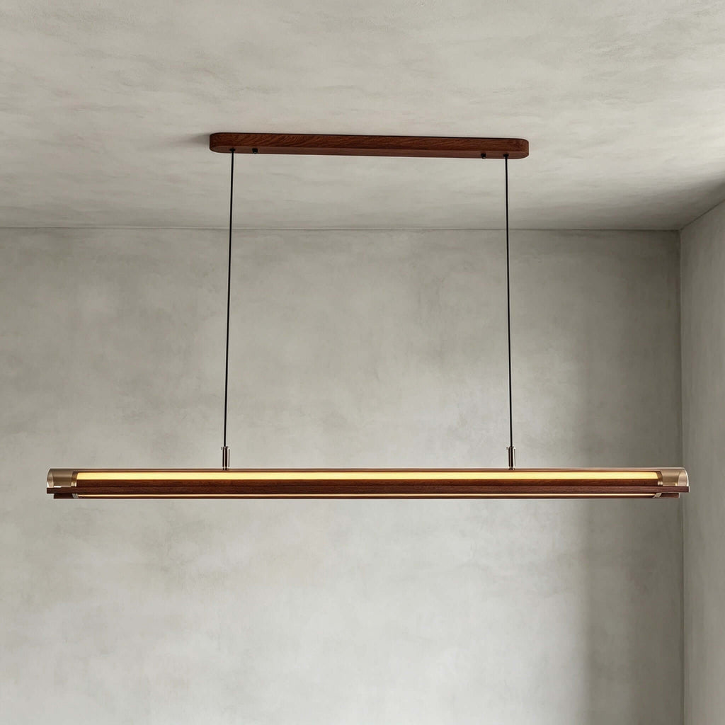 A Clérachic linear pendant hangs from a walnut canopy on two cables, lit warm, in front of a grey plaster wall.