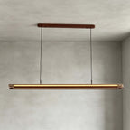 A Clérachic linear pendant hangs from a walnut canopy on two cables, lit warm, in front of a grey plaster wall.