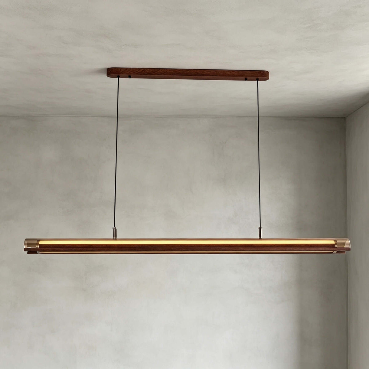 A Clérachic linear pendant hangs from a walnut canopy on two cables, lit warm, in front of a grey plaster wall.