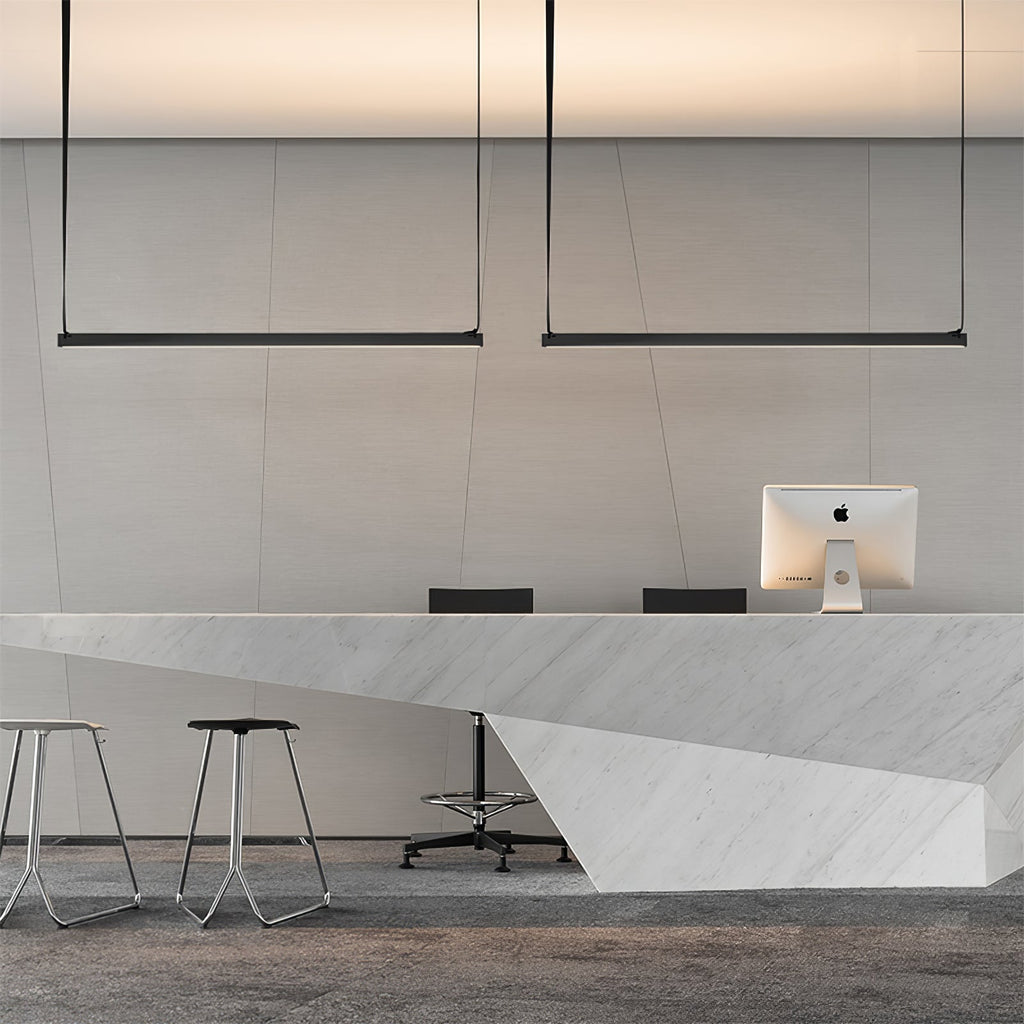 Two Clérachic linear pendant lights hang from straps above a white stone reception counter with stools and a desktop computer.