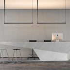 Two Clérachic linear pendant lights hang from straps above a white stone reception counter with stools and a desktop computer.