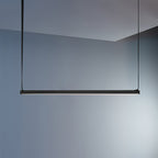 A black bar of this Clérachic linear pendant light hangs on its straps against a soft blue-grey wall, with light along its underside.