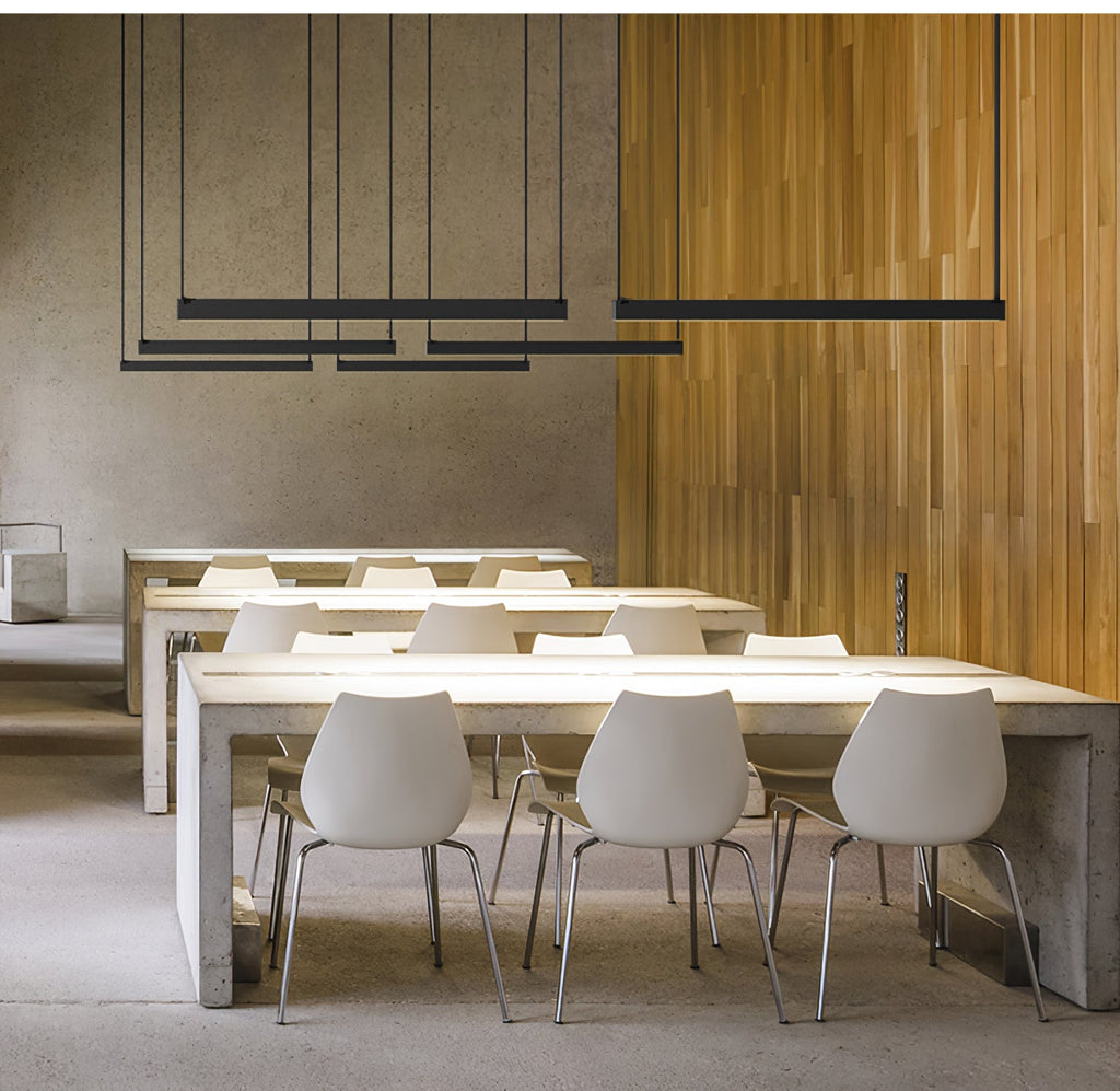 Several black bars from this Clérachic linear pendant light hang at varied heights over long white tables in a pale concrete hall.