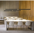 Several black bars from this Clérachic linear pendant light hang at varied heights over long white tables in a pale concrete hall.