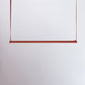 A red bar of this Clérachic linear pendant light hangs from two red straps against a pale lilac-grey wall.