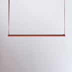 A red bar of this Clérachic linear pendant light hangs from two red straps against a pale lilac-grey wall.
