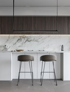 Above a marble splashback and white island with grey stools, this Clérachic linear pendant light hangs as a slim black bar.