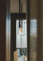Looking up at one strap and bar of this Clérachic linear pendant light, its orange-lit underside showing against a dark doorway.