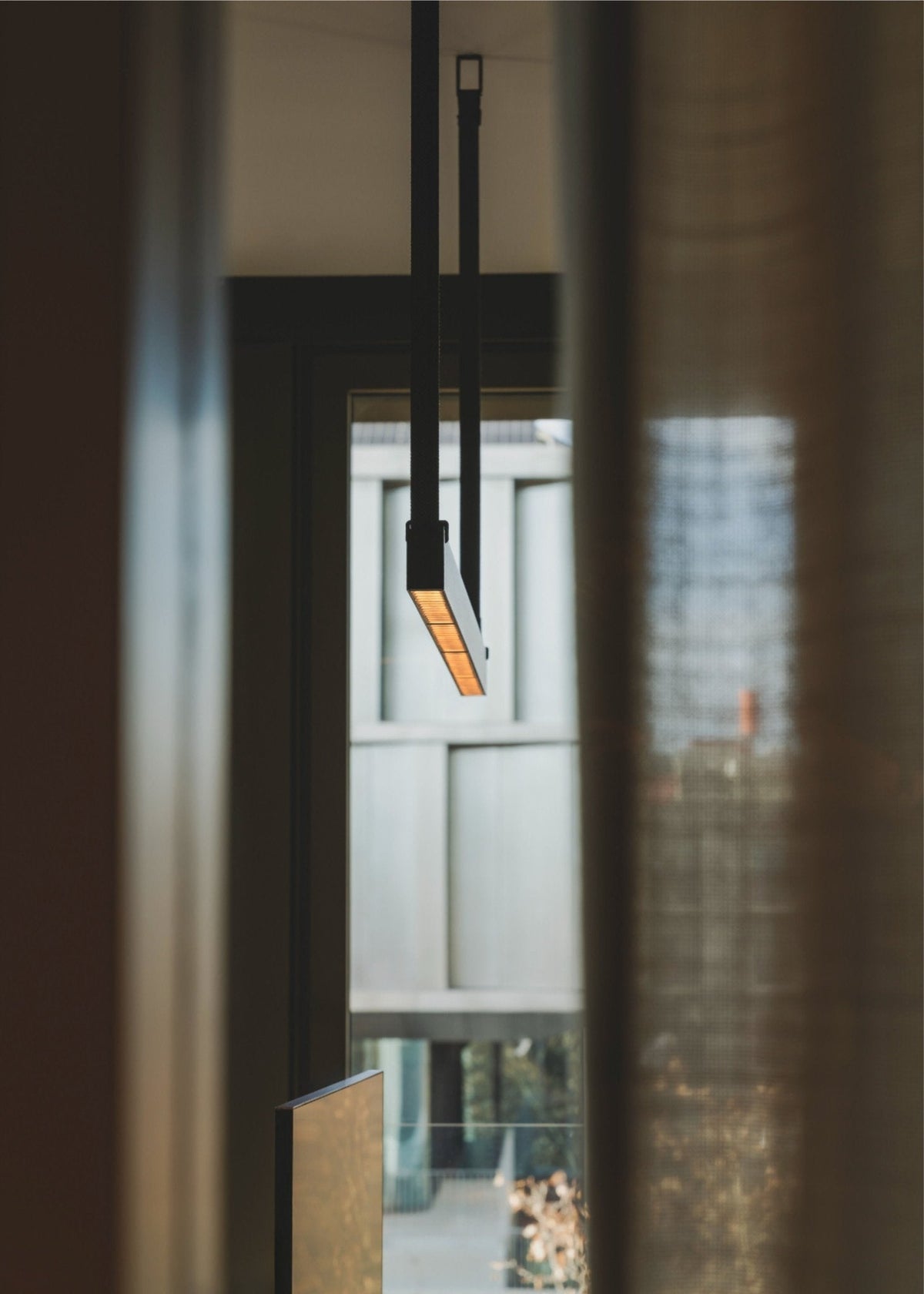 Looking up at one strap and bar of this Clérachic linear pendant light, its orange-lit underside showing against a dark doorway.