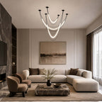 Black Clérachic pendant of three lit curved tubes over a sectional sofa and a marble coffee table in a neutral living room.