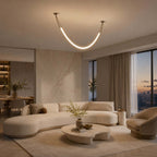 A single gold Clérachic curved light tube hung from two canopies over a curved cream sofa, with a city dusk view in the window.