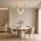 Single gold Clérachic curved tube light above a round stone-based dining table with four cream chairs on gold legs.