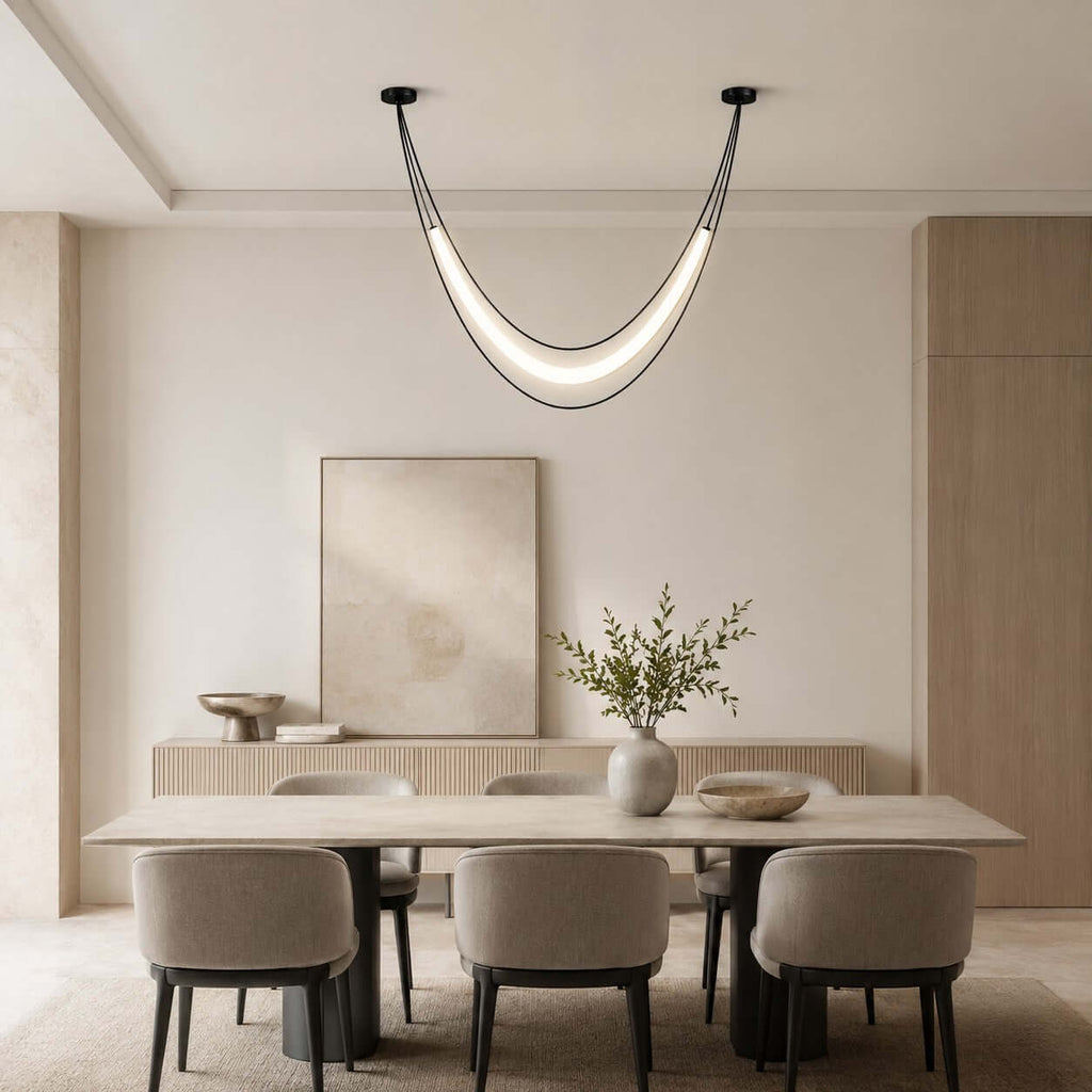 Black Clérachic curved pendant with a lit white tube over a long dining table, with a pale artwork on the sideboard behind.