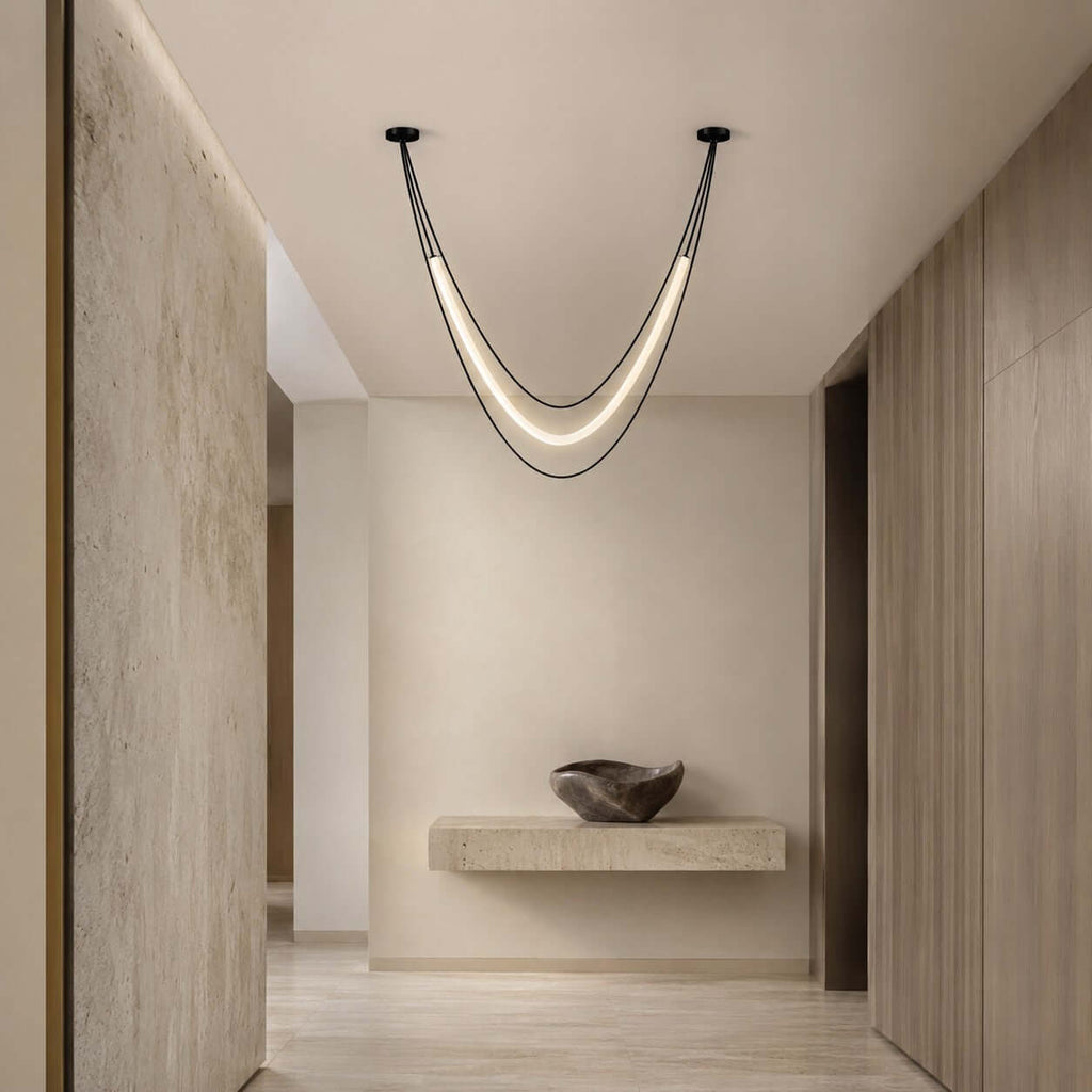 Black Clérachic pendant with a softly glowing tube, hung at a hallway junction before a floating stone shelf and a stone bowl.