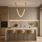 Clérachic gold pendant, a lit curved tube on fine cords, over a ribbed-front kitchen island with brass-legged stools.