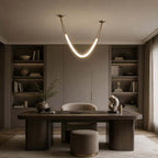 Clérachic curved light tube in gold and white over a dark desk in a library-style room, with shelves and a sculpture beside.
