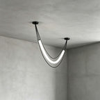 Black Clérachic pendant with one lit white curved tube and two cords, seen close in the corner of a concrete-look room.