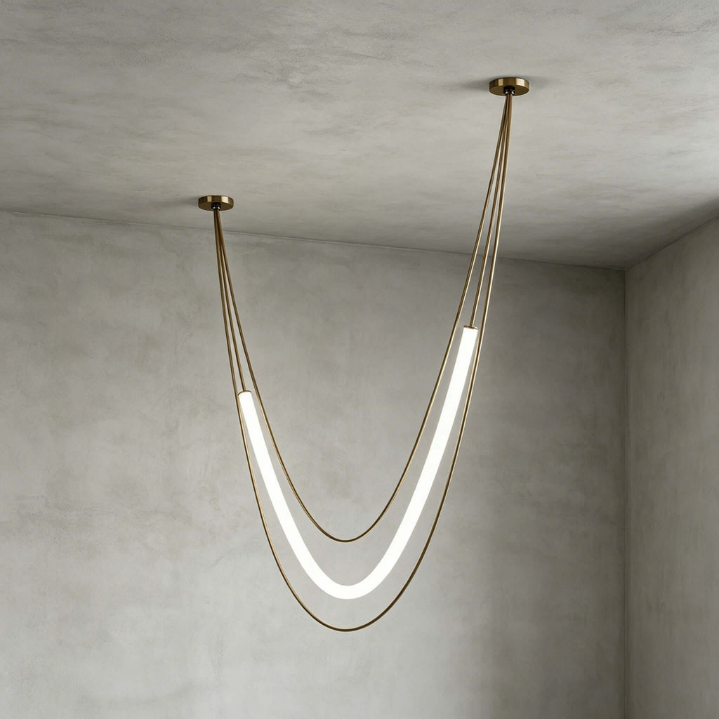 Gold Clérachic pendant with a tall lit tube on slim gold cords, canopies at different heights, in a grey plaster room corner.