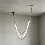 Gold Clérachic pendant with a tall lit tube on slim gold cords, canopies at different heights, in a grey plaster room corner.