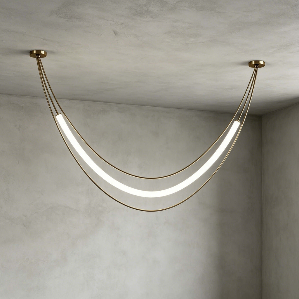 Gold Clérachic pendant in a wide shallow curve, its white tube glowing between two ceiling canopies on a grey plaster wall.
