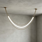 Gold Clérachic pendant in a wide shallow curve, its white tube glowing between two ceiling canopies on a grey plaster wall.