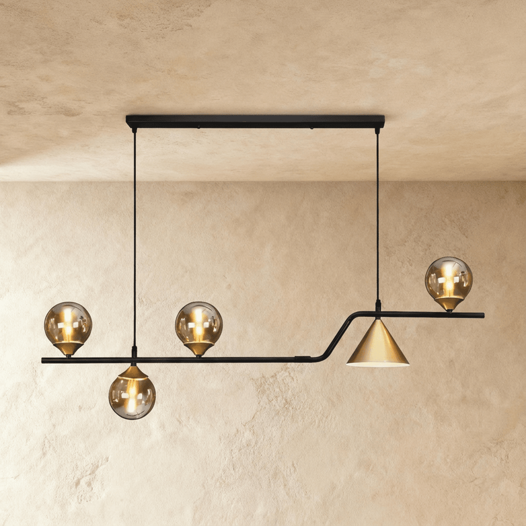 Clérachic linear pendant with four tinted glass globes and a gold cone on a bent black bar, in a studio on a limewash wall.