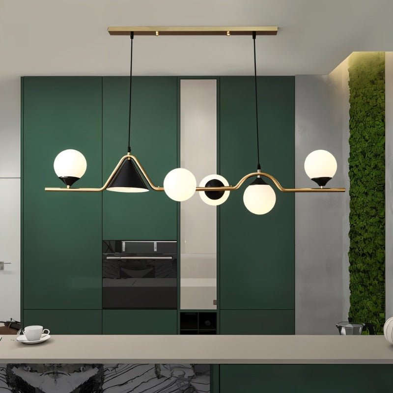 Clérachic gold and black linear pendant with milk white globes, hung in a room above a kitchen counter and green cabinets.