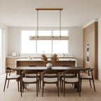 A slim Clérachic linear pendant hangs in front of a large window, above a dark dining table with wooden chairs.