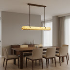A lit Clérachic linear pendant hangs above a long walnut dining table with cream upholstered chairs and tall curtains.