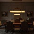 A lit Clérachic linear pendant hangs over a dark dining table, below a gilt mirror and landscape paintings.