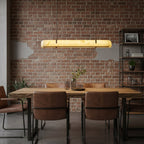 A lit Clérachic linear pendant hangs against a brick wall, above a rustic wooden table with leather chairs.