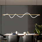 Brass Clérachic linear pendant with a glowing wave, above a wood dining table in front of a grey slatted wall.