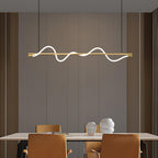 Lit brass Clérachic linear pendant above a pale table with tan chairs, against a warm taupe panelled wall.