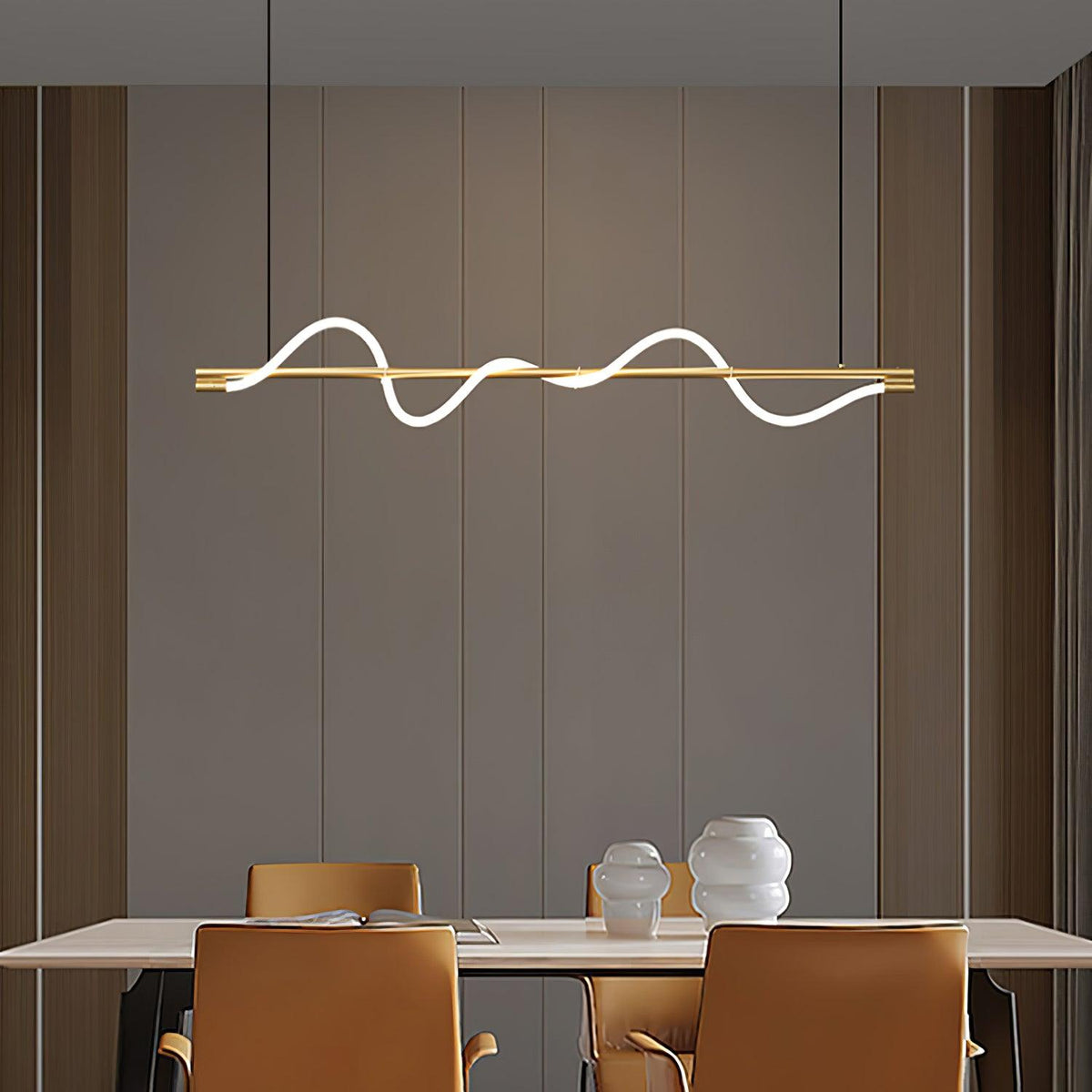 Lit brass Clérachic linear pendant above a pale table with tan chairs, against a warm taupe panelled wall.