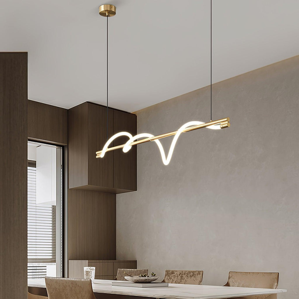 Angled view of a brass Clérachic linear pendant over a dining table, beside timber cabinets and a window.
