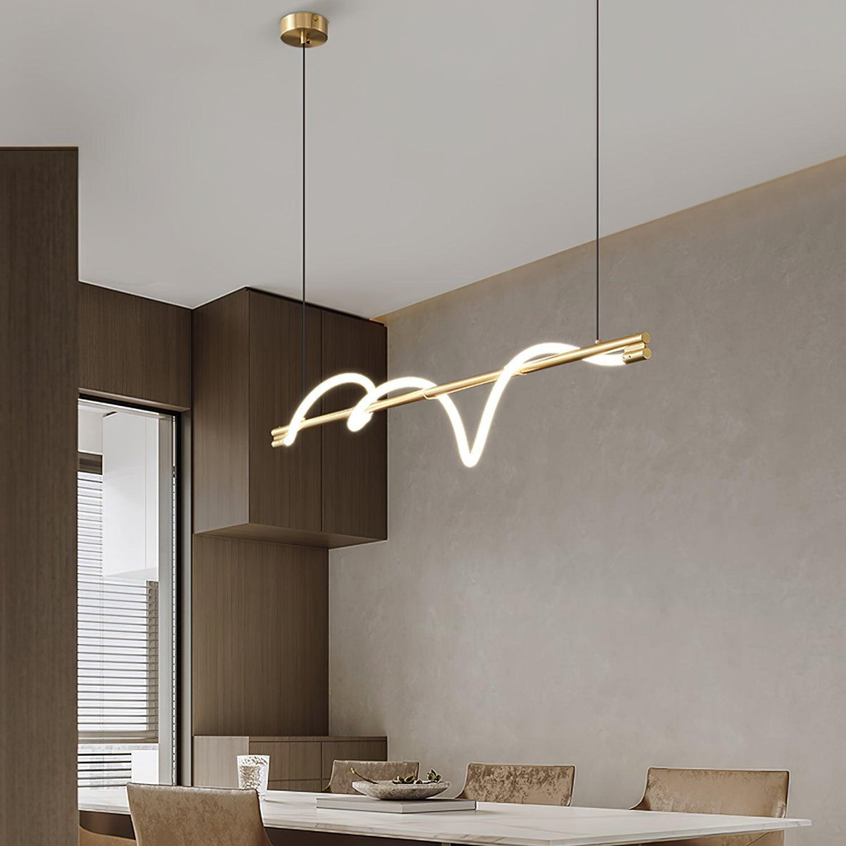 Angled view of a brass Clérachic linear pendant over a dining table, beside timber cabinets and a window.