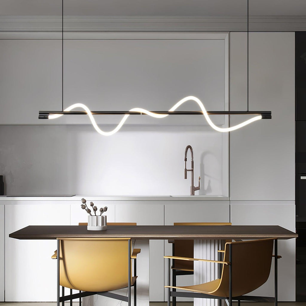 A black Clérachic linear pendant with one glowing wave hangs above a dark table with mustard chairs in a pale kitchen.