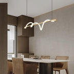 Slanted brass Clérachic linear pendant above a pale stone table and tan chairs, with wooden cabinets beside it.