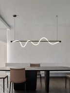 Side-angled view of a black Clérachic linear pendant lit above a dark table with a tan leather chair.